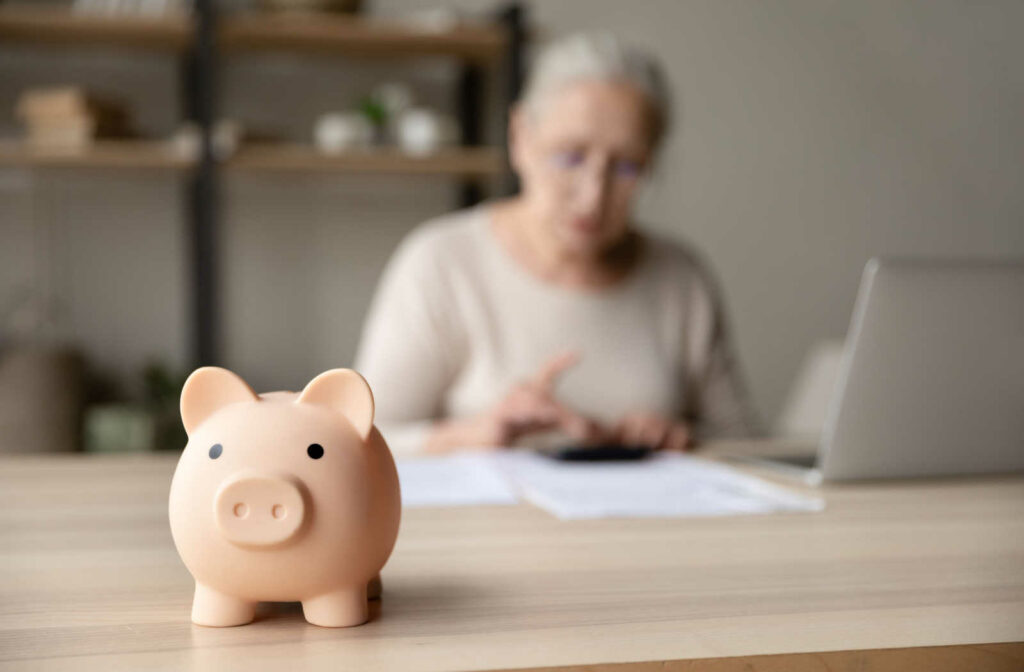 An out of focus mature woman sitting at a table with a laptop and papers with an in focus piggy bank on the table in front of her.