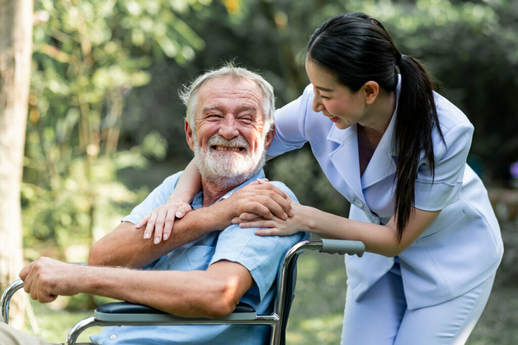 A female nurse is assisting an elderly man with Alzheimer's on a wheelchair to go outdoors in the garden.