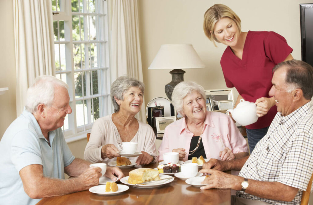 Group of seniors sitting around a table enjoying afternoon tea smiling and chatting with each other with their nurse in a senior living facility.