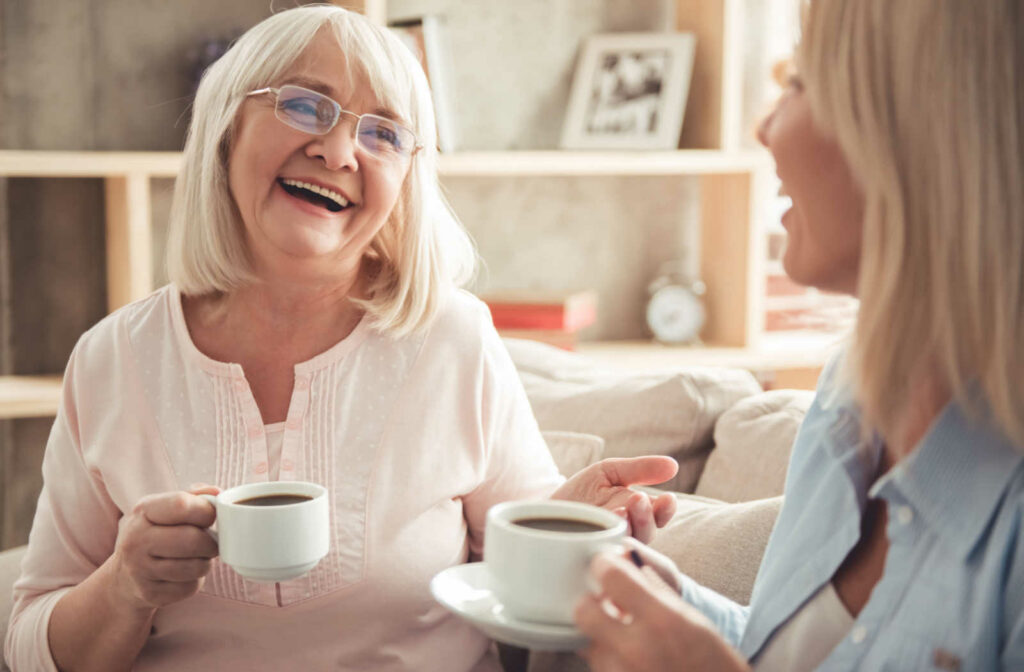 A senior woman and her daughter sitting on a couch smiling and talking to each other while holding a cup of coffee.