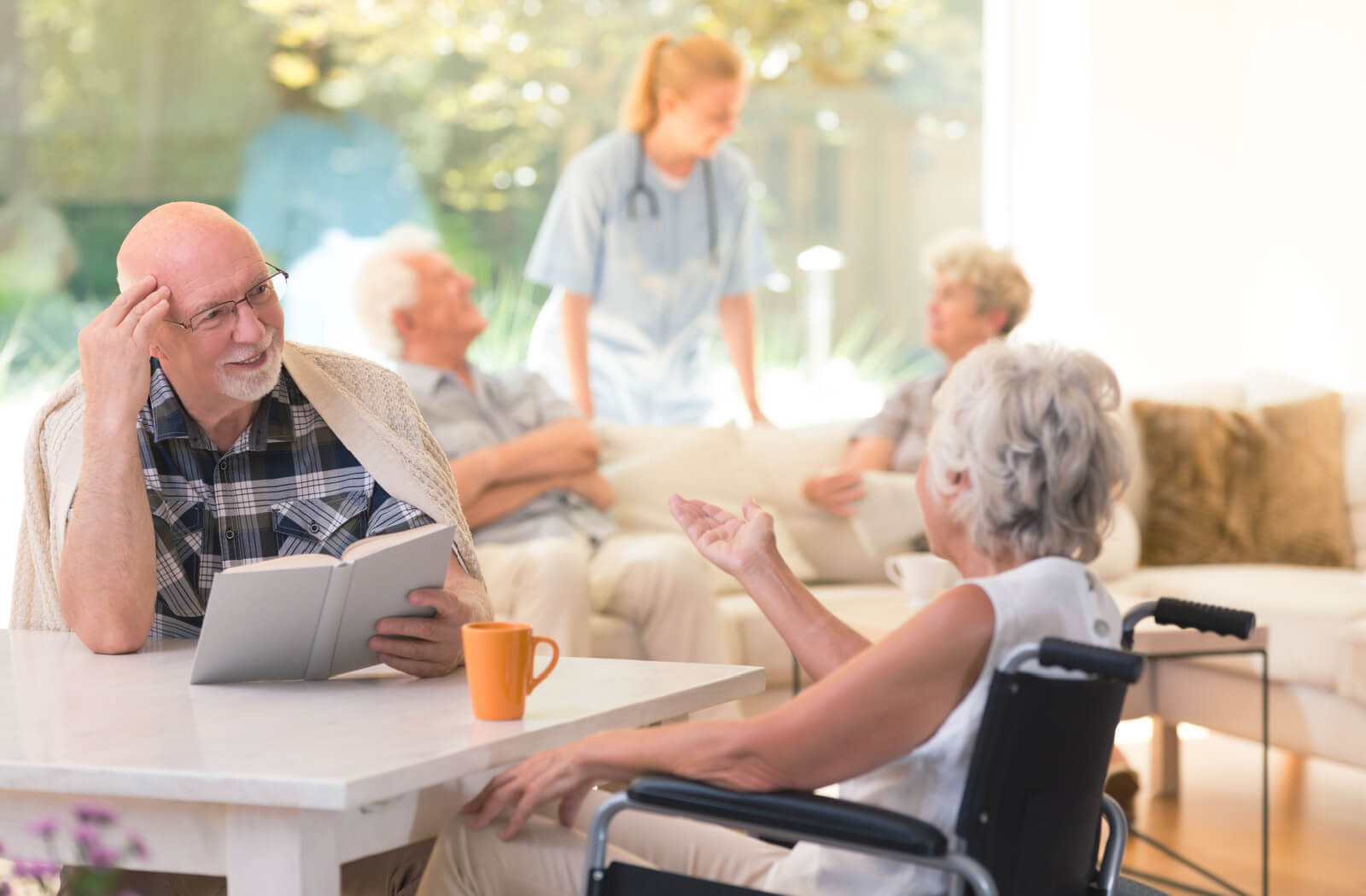 A group of seniors sitting socializing indoors engaged in various activities such as reading and conversing with each other.