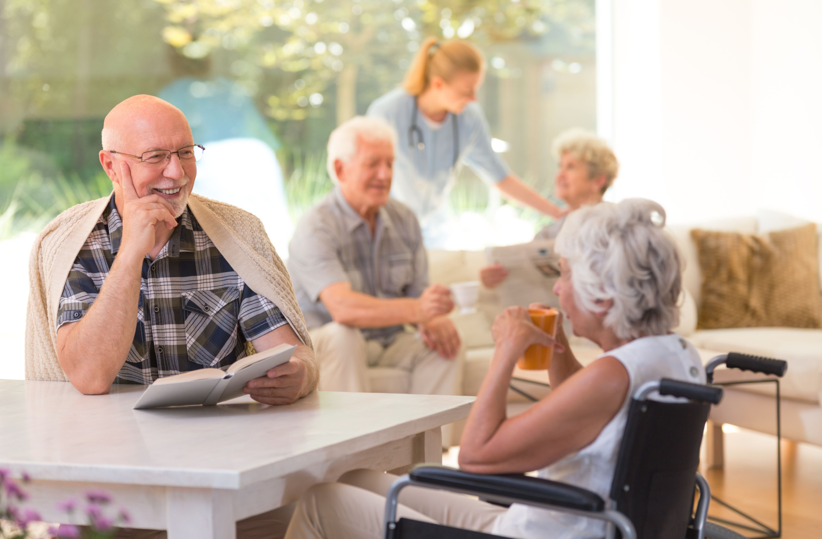 A senior man sitting at a table holding a book and engaged in conversation with an elderly woman in a wheelchair.