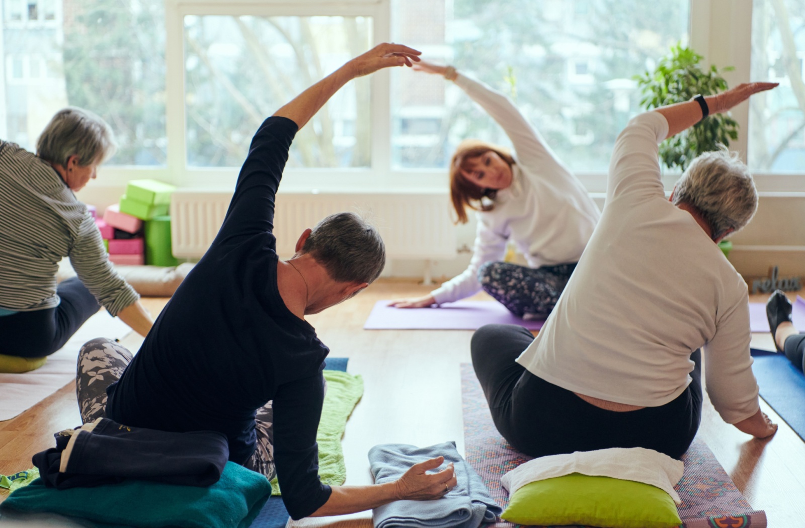 A group of seniors are practicing light yoga in a brightly lit studio, following a teacher on brightly coloured yoga mats.