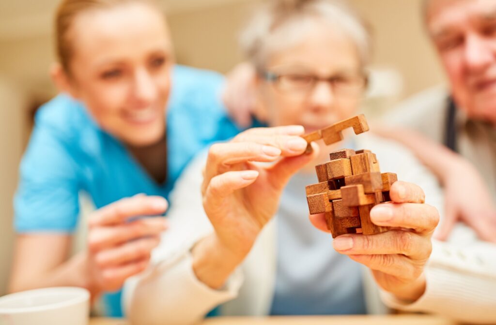 An older person with dementia puts together a puzzle. A friend and caregiver are seen behind her.