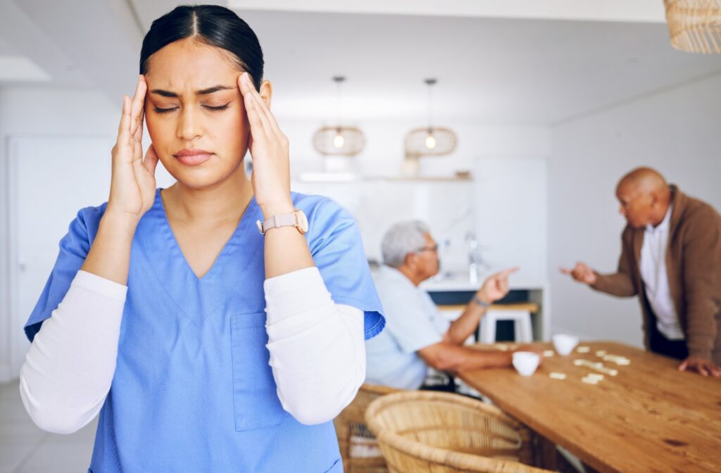 A caregiver massages her temples in exhaustion as two older adults argue and gesture at each other in the background.