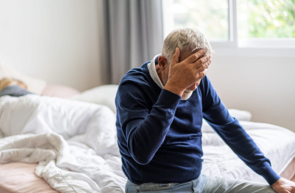 An older adult in assisted living feeling disoriented and holding their head while seated on a bed.