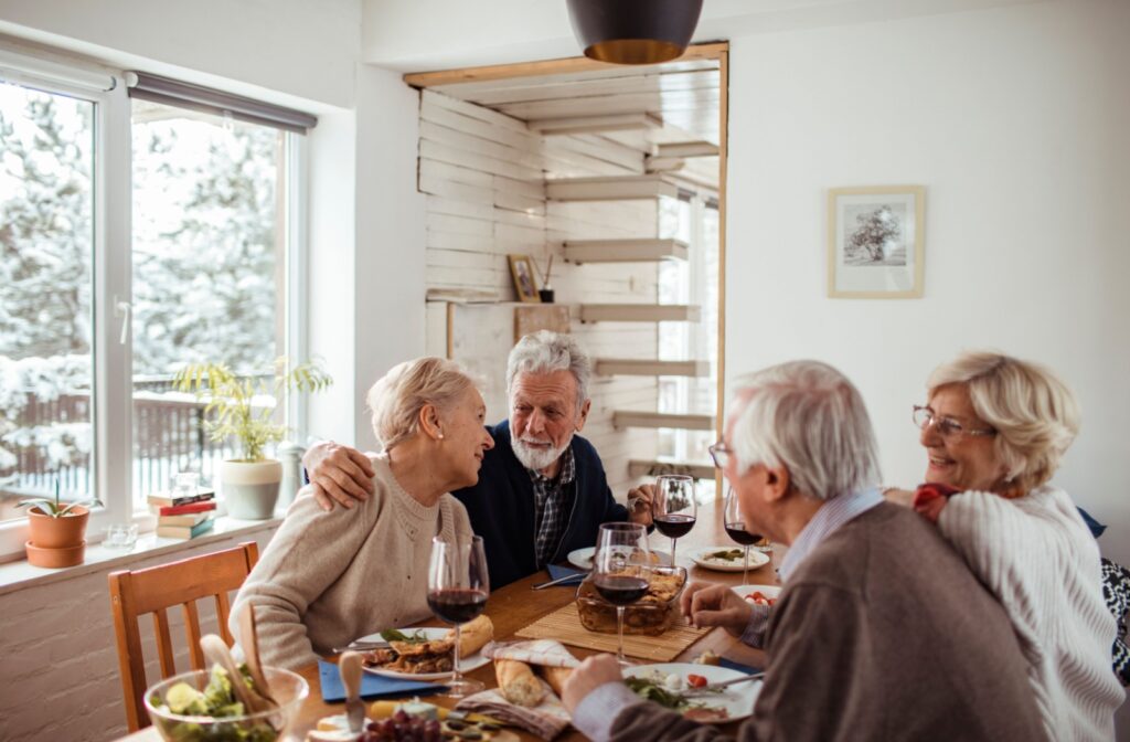 4 older adults laugh over a shared meal in their home in senior living while it snows outside.
