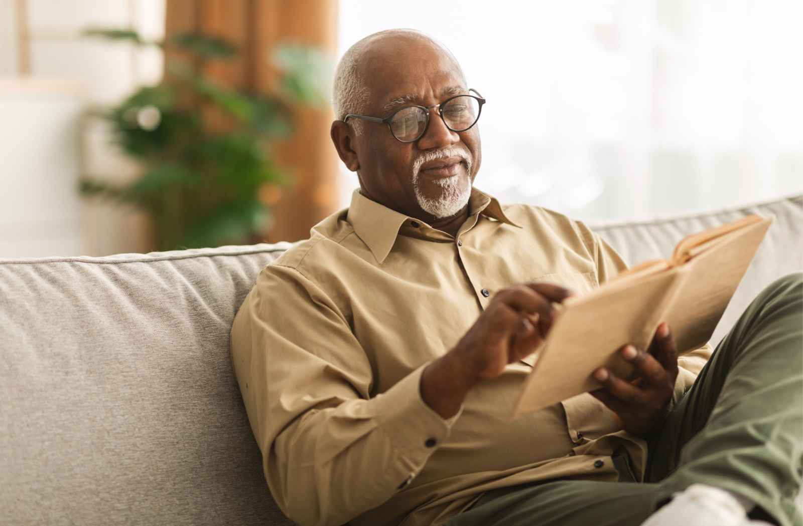 A senior man sitting on a couch wearing eyeglasses and reading a book.