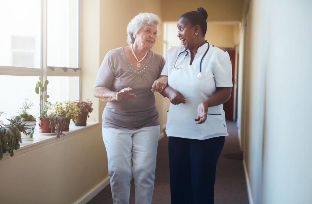 A caregiver walks down a hallway arm-in-arm with a smiling senior beside windows and potted plants